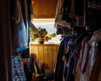 A colour photo of a wardrobe, clothes hanging on both sides. At the end is a small window with a fairly large and bulbous potted cactus.
